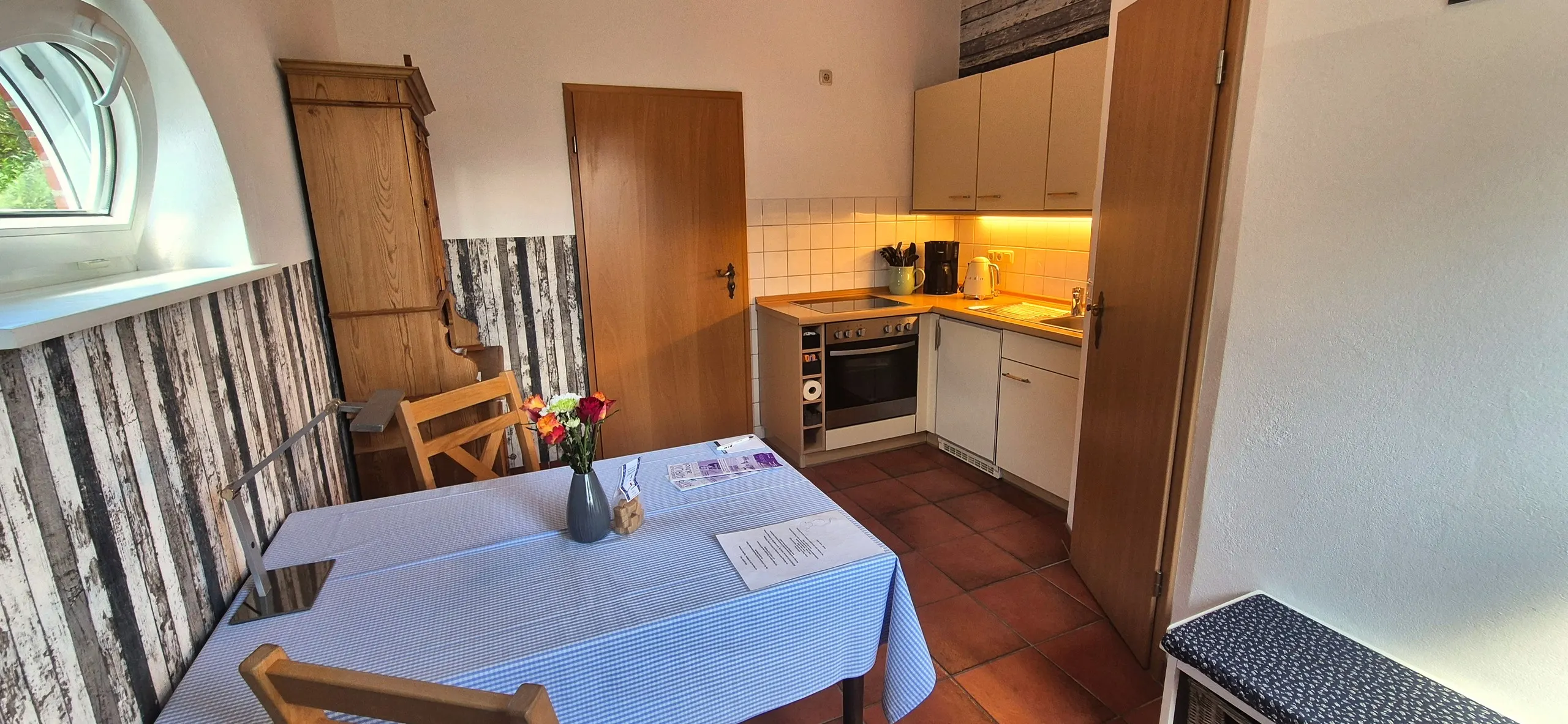 Tanzmaus – Kleine Wohnküche mit Esstisch mit blau-weiß karierter Tischdecke, Blumenstrauß und Infomappe im Vordergrund, dahinter …