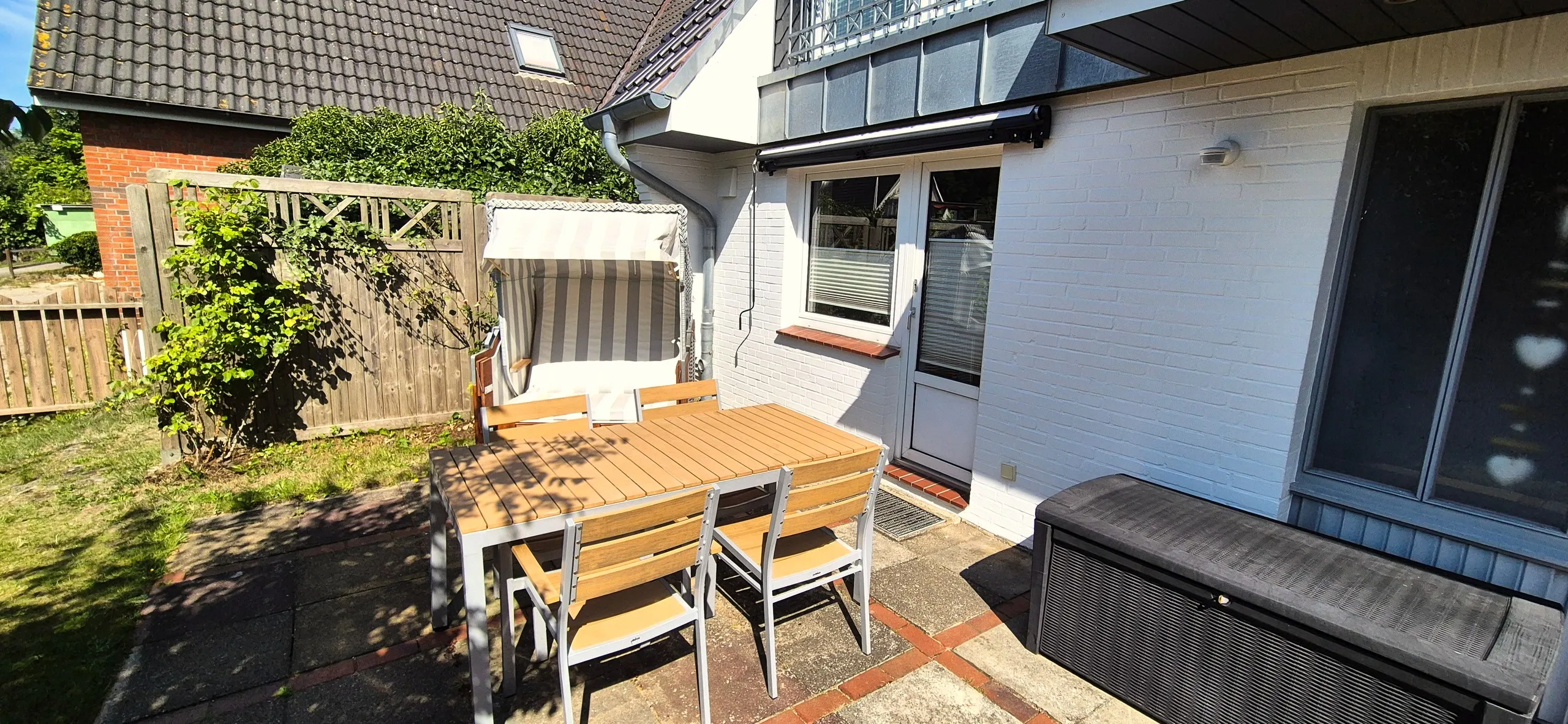 Butt – Terrasse mit Holztisch, vier Stühlen und einem Strandkorb vor der weißen Hausfassade.