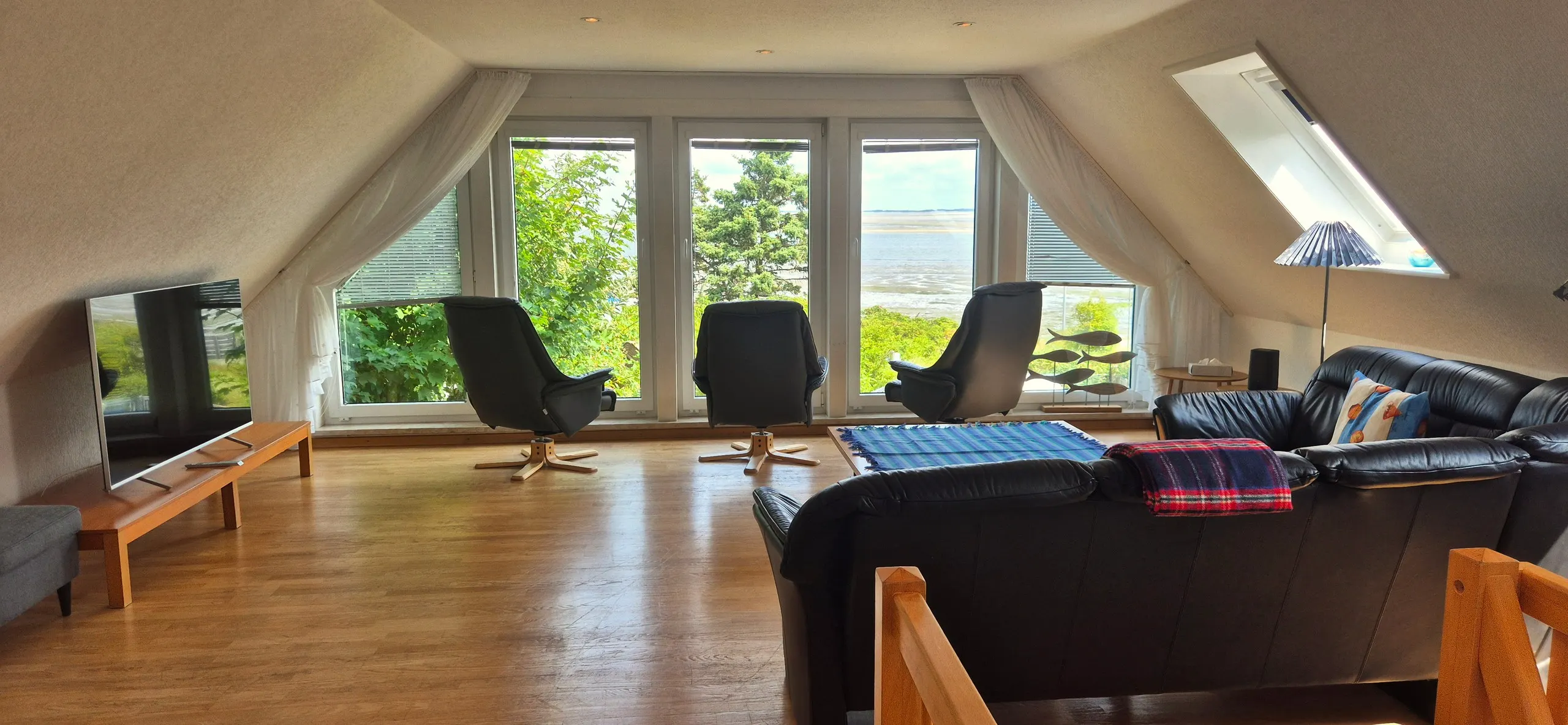 Küstenhaus – Weiträumiges Wohnzimmer unter dem Dach mit schwarzer Leder-Sofalandschaft im Vordergrund, davor ein Couchtisch mit …