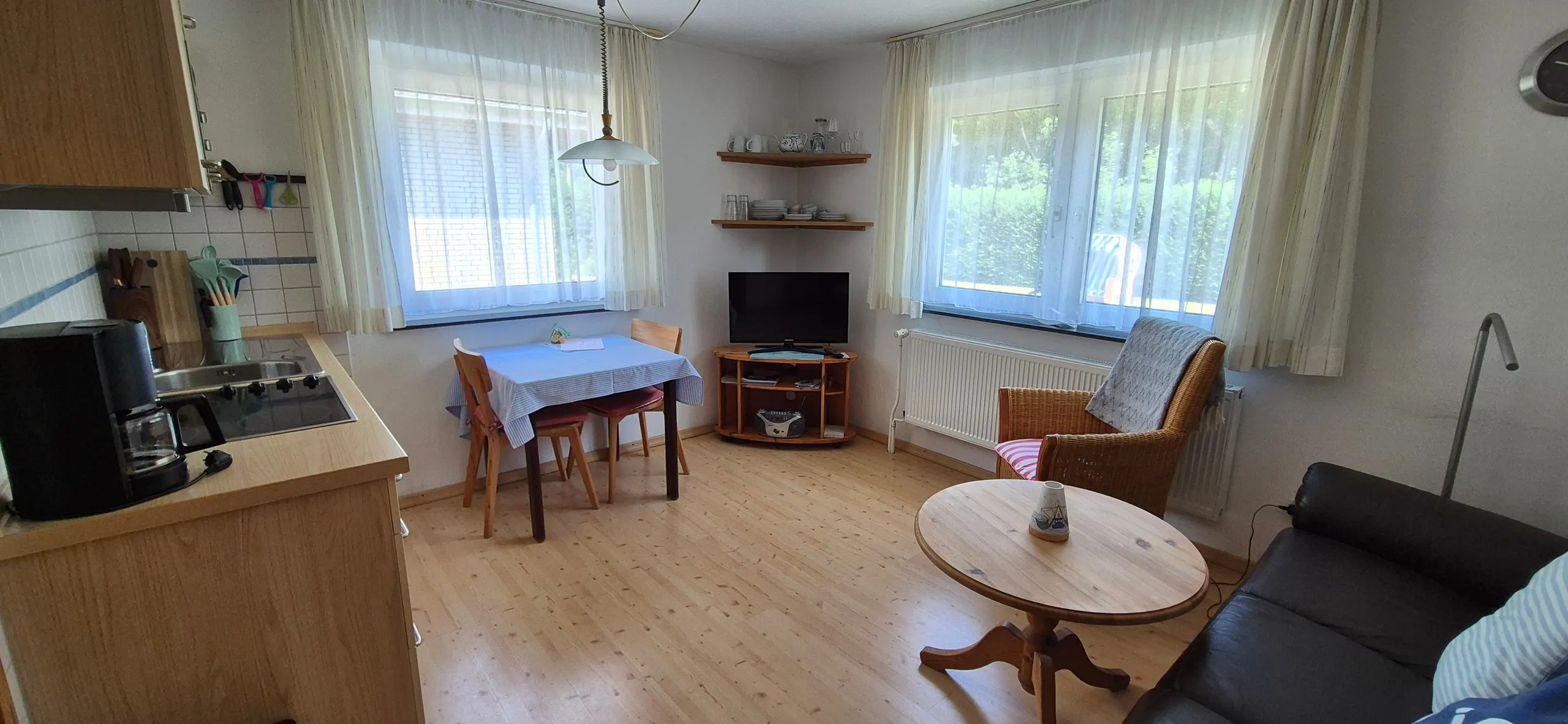 Enzian – Wohn-Esszimmer mit kleinem Esstisch, blau-weiß gestreifter Tischdecke und zwei Holzstühlen vor einem Fenster mit hellen …