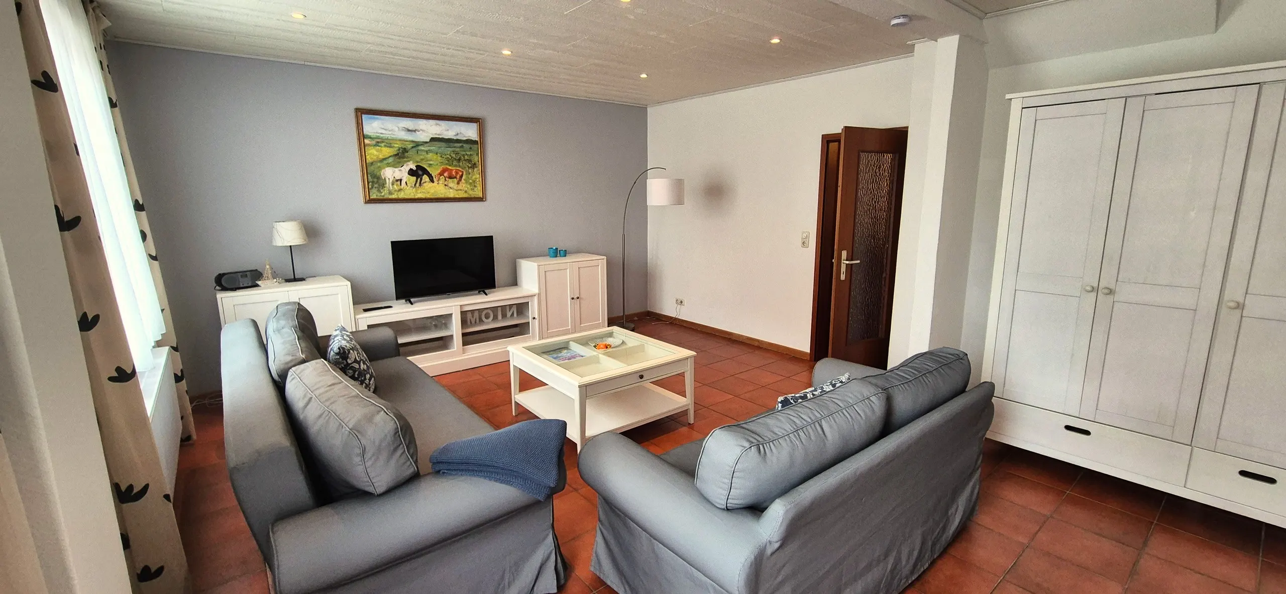 Spitzmaus – Wohnzimmer mit zwei grauen Sofas um einen weißen Couchtisch, Flachbildfernseher auf einem TV-Board und einem großen …