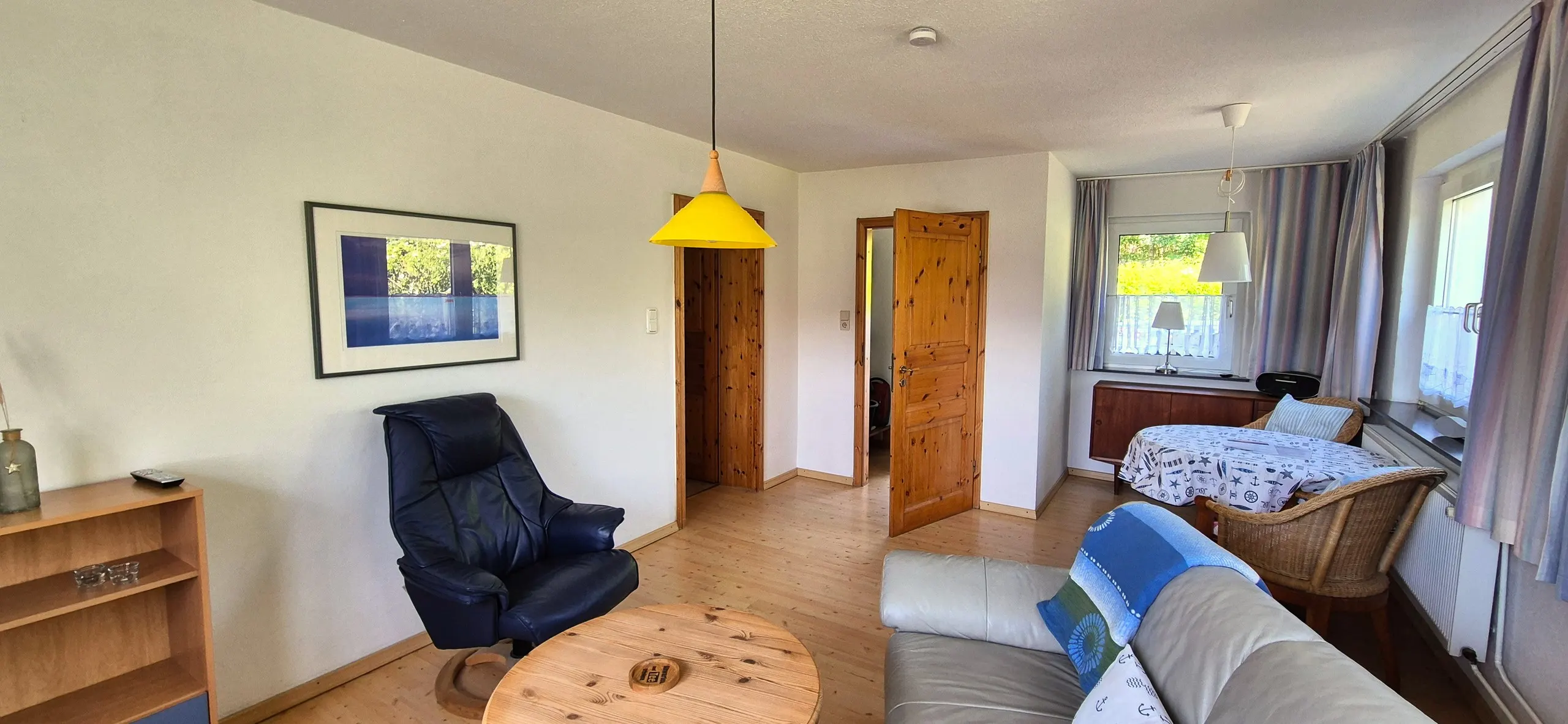 Grasnelke – Offener Wohn-Essbereich mit dunklem Lederrelaxsessel und rundem Kiefernholz-Couchtisch im Vordergrund, an der Wand …