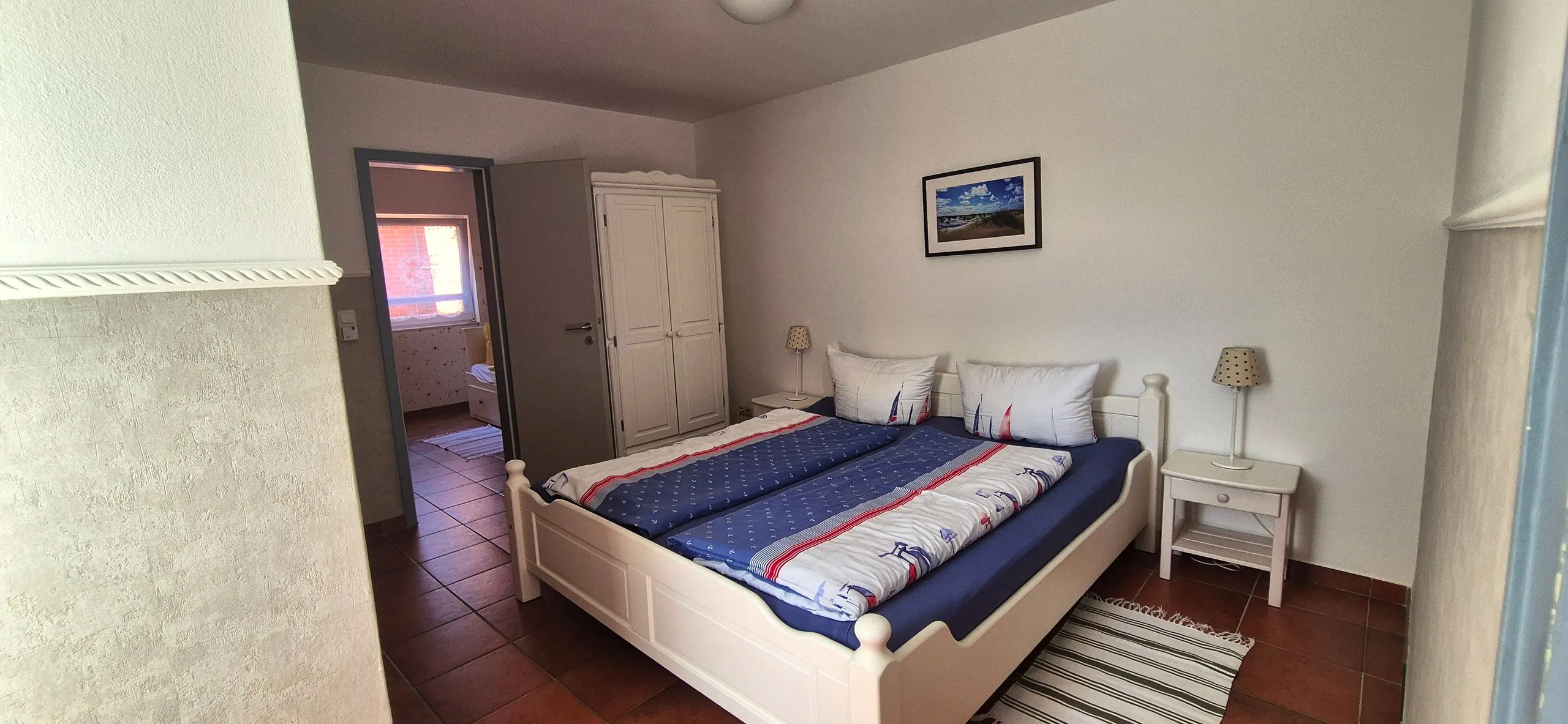 Feldmaus – Geräumiges Schlafzimmer mit weißem Doppelbett im Landhausstil und maritimer Bettwäsche in Blau-Weiß mit Anker- und …