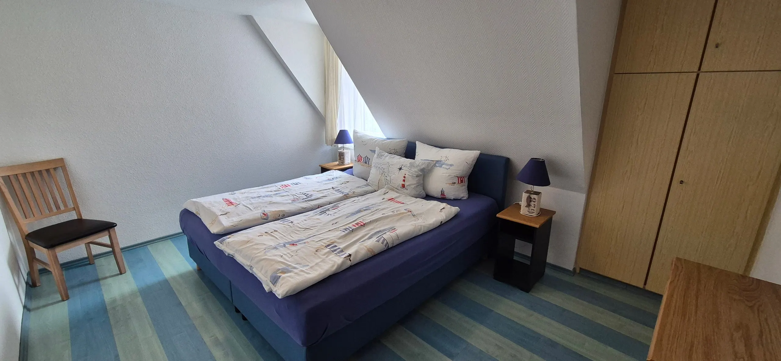 Dünenrose – Schlafzimmer unter dem Dach mit dunkelblauem gepolstertem Doppelbett und maritimer Bettwäsche mit Leuchtturm-Motiven.