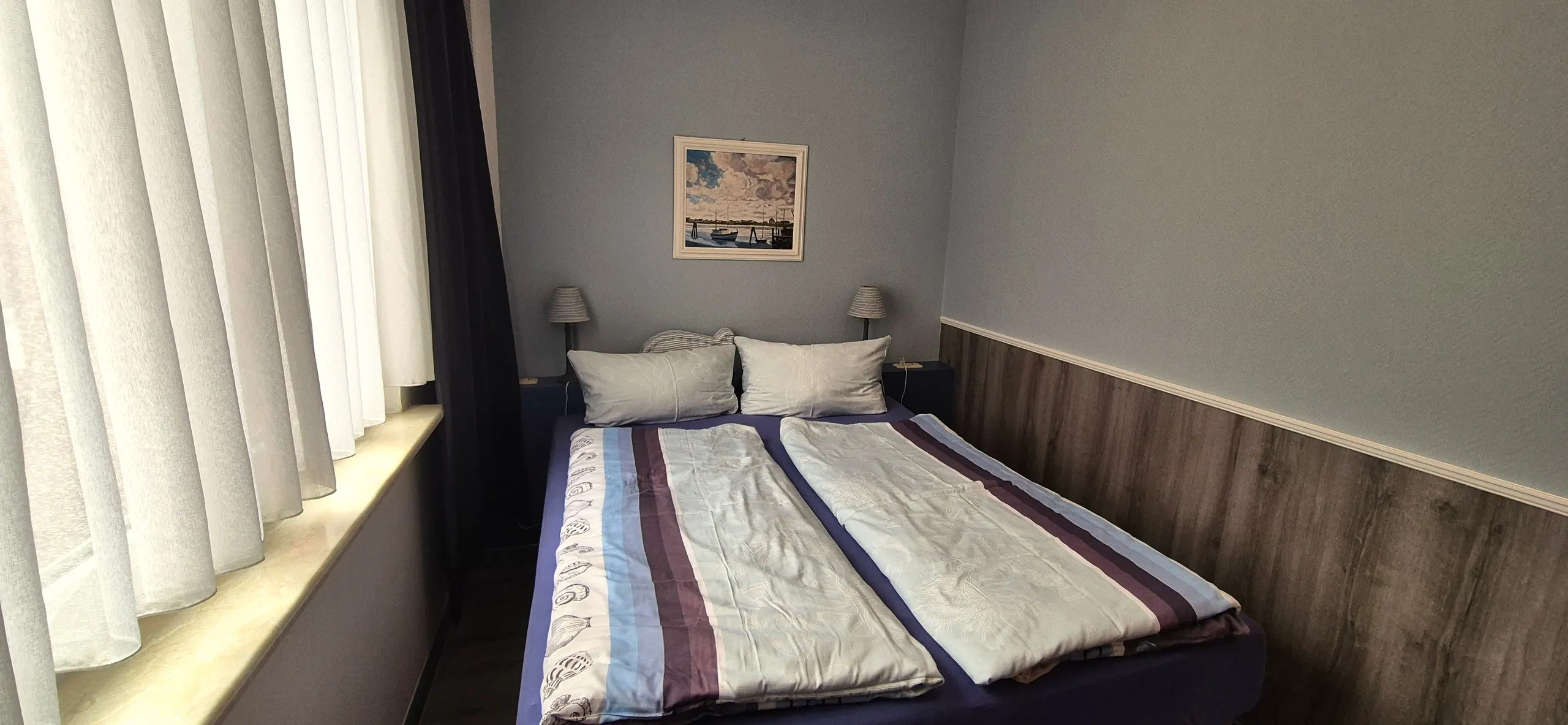 Spitzmaus – Schlafzimmer mit bezogenem Doppelbett, zwei Nachttischleuchten und einem Segelboot-Gemälde über dem Kopfteil.