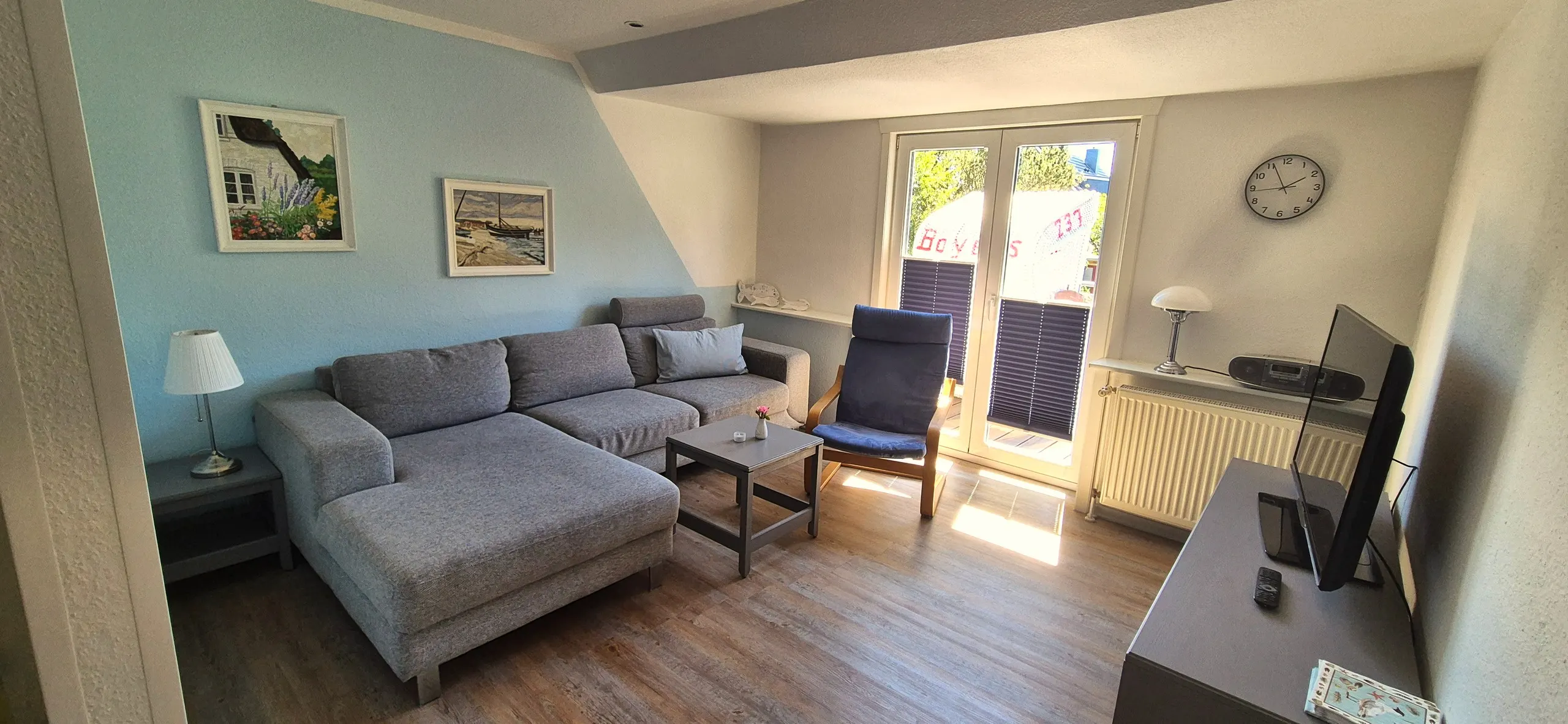 Regenpfeifer – Wohnzimmer mit großer grauer Ecksofalandschaft und passendem Sessel im Skandi-Stil, dazwischen ein kleiner Couchtisch …