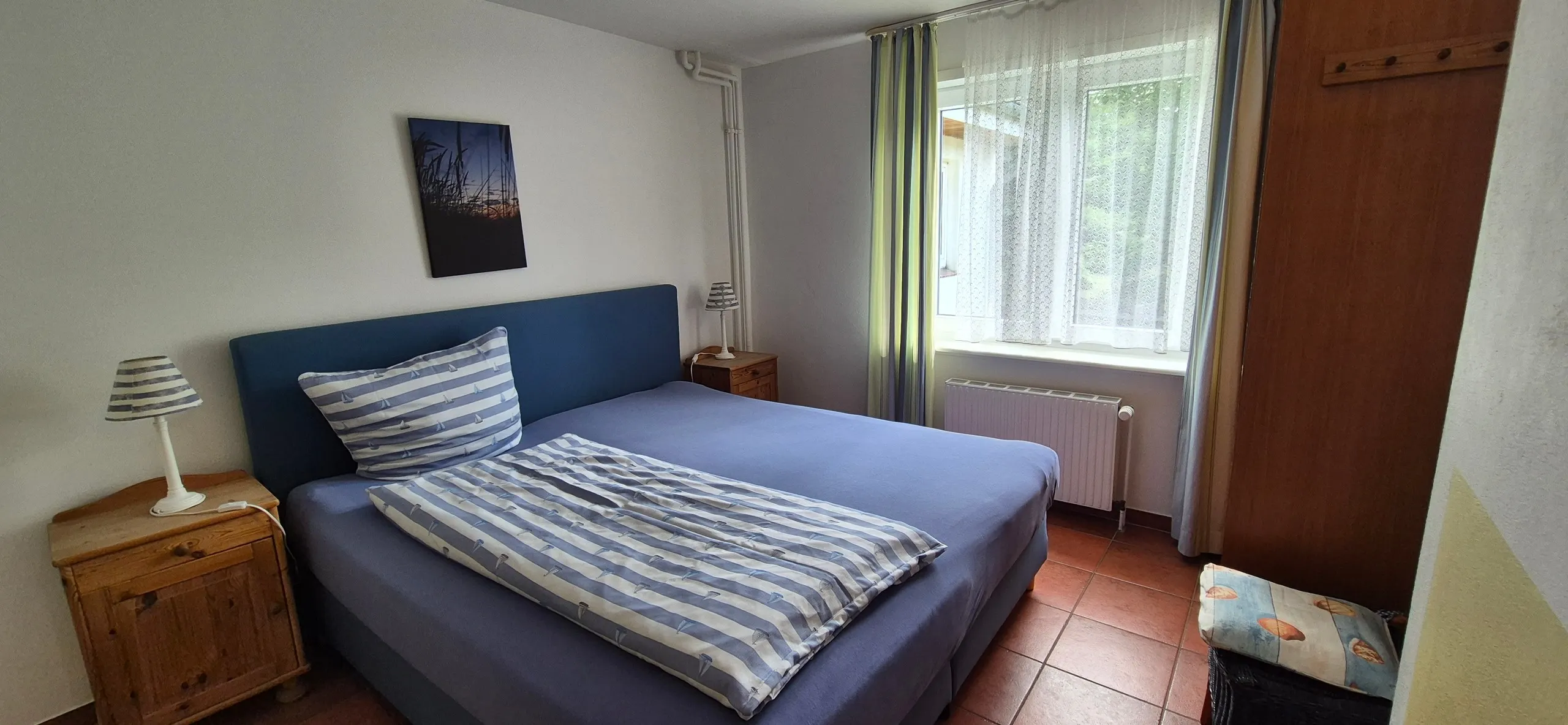 Strandflieder – Schlafzimmer mit dunkelblauem, gepolstertem Doppelbett und grau-weiß gestreifter Bettwäsche.