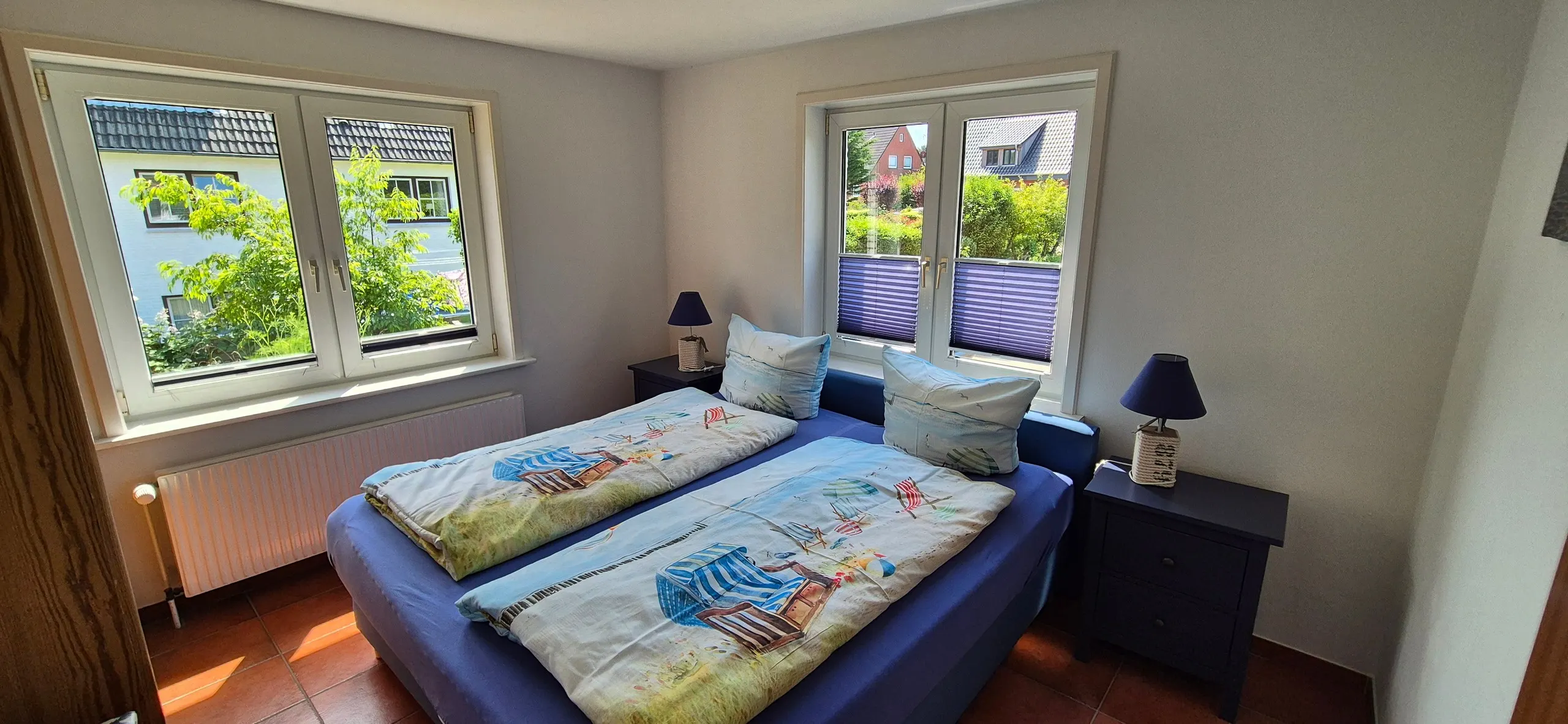 Sonnentau – Helles Schlafzimmer mit breitem Doppelbett in Blau, bezogen mit maritimer Bettwäsche mit Strandkorb-Motiven.