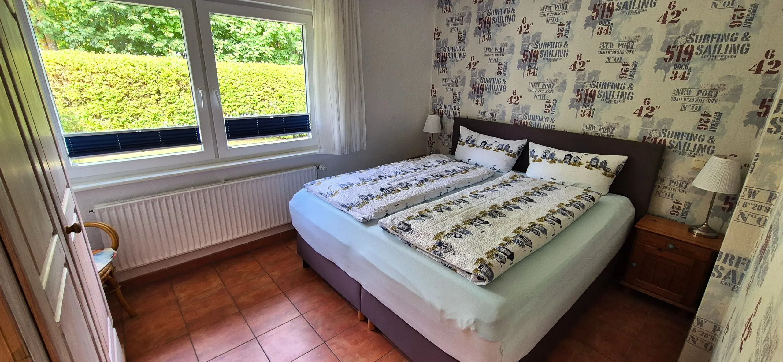 Austernfischer – Doppelbett vor einer Tapete mit maritimen Segel-Motiven, davor ein Fenster mit Blick auf eine grüne Hecke.