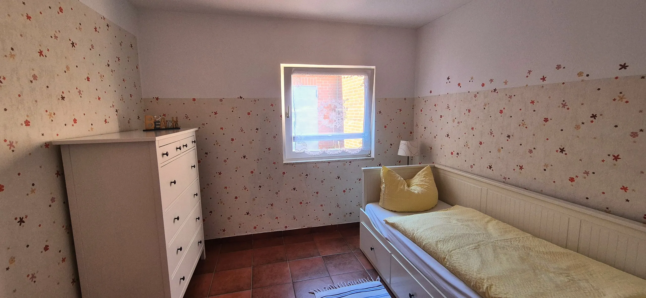 Feldmaus – Kleines Schlafzimmer mit Einzelbett und gelber Bettwäsche, die Wände sind mit einer zart geblümten Tapete gestaltet.