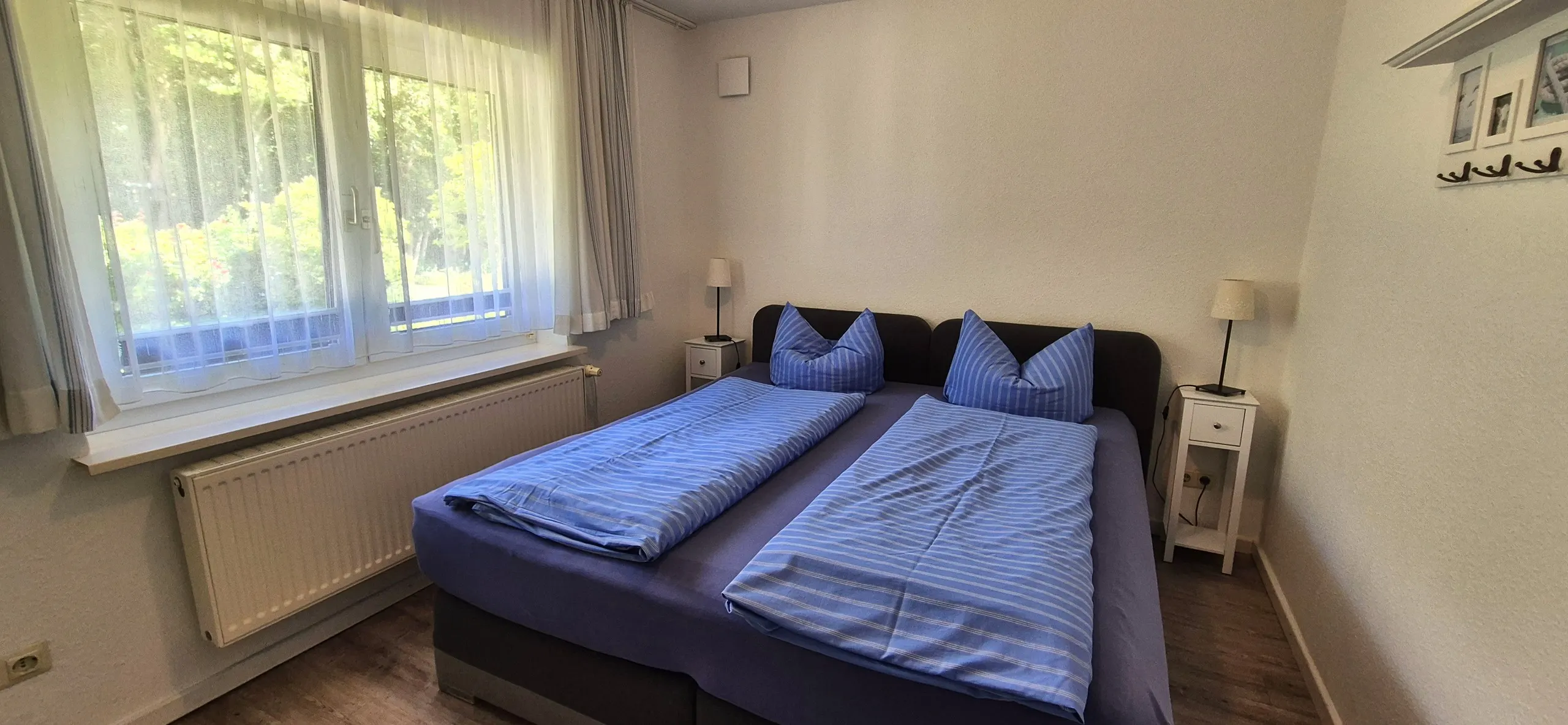 Krabbe – Schlafzimmer mit Doppelbett, gestreifter Bettwäsche in Blau, zwei weißen Nachttischen mit Lampen und großem Fenster mit …