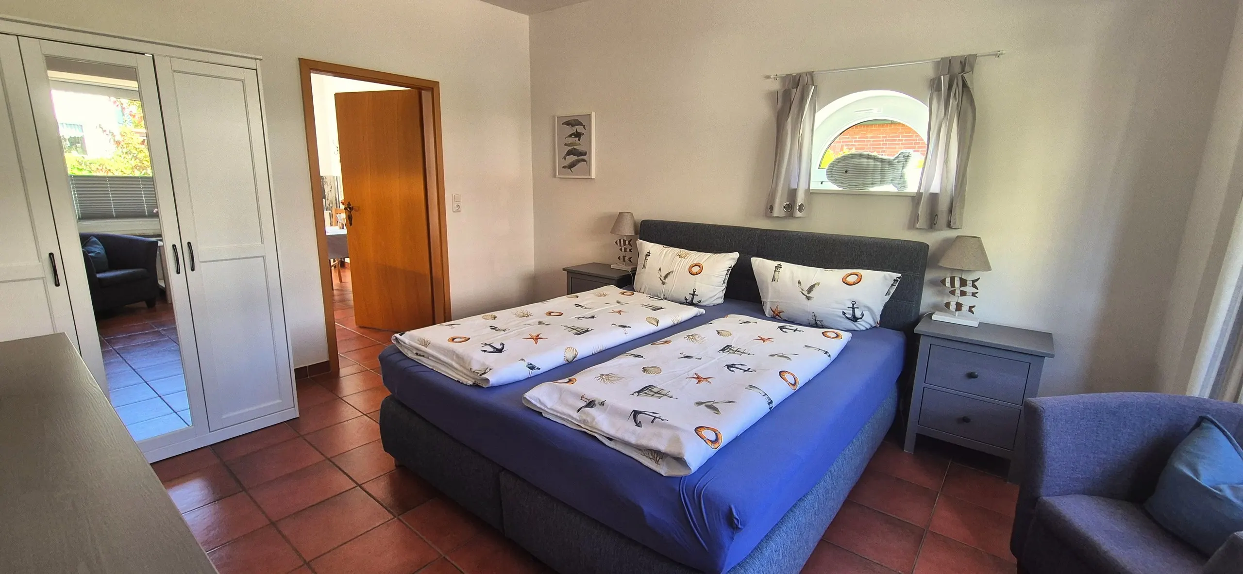 Tanzmaus – Zweites Schlafzimmer mit Doppelbett mit maritimer Bettwäsche, davor ein Spiegelschrank links und ein halbrundes Fenster …