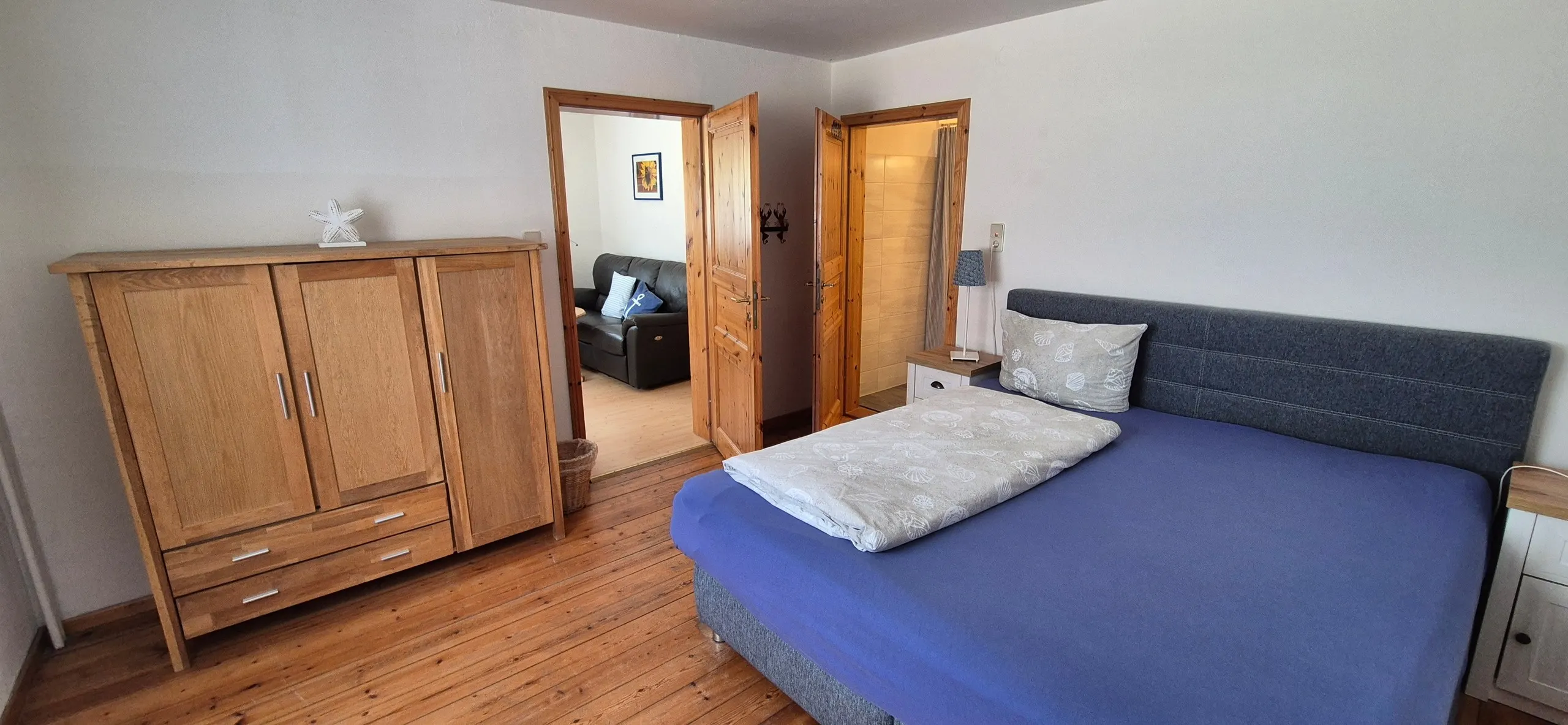 Enzian – Schlafzimmer mit Doppelbett, dunkelblauem Bettbezug und maritimen Kissen mit Muschelmotiv vor gepolstertem, …