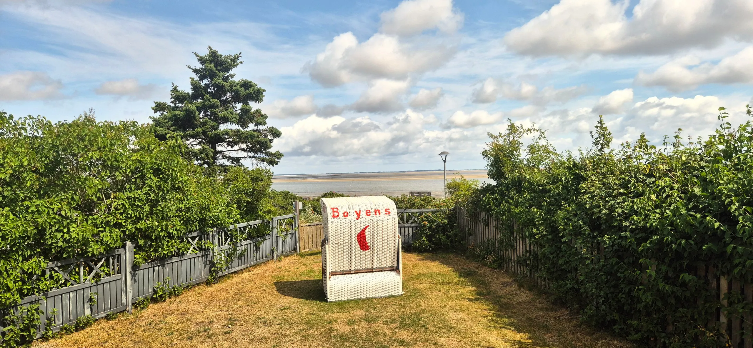 Küstenhaus – Gepflegter, von Hecken und Holzzäunen eingefasster Garten mit weißem Strandkorb mit rotem Segel-Motiv und der …