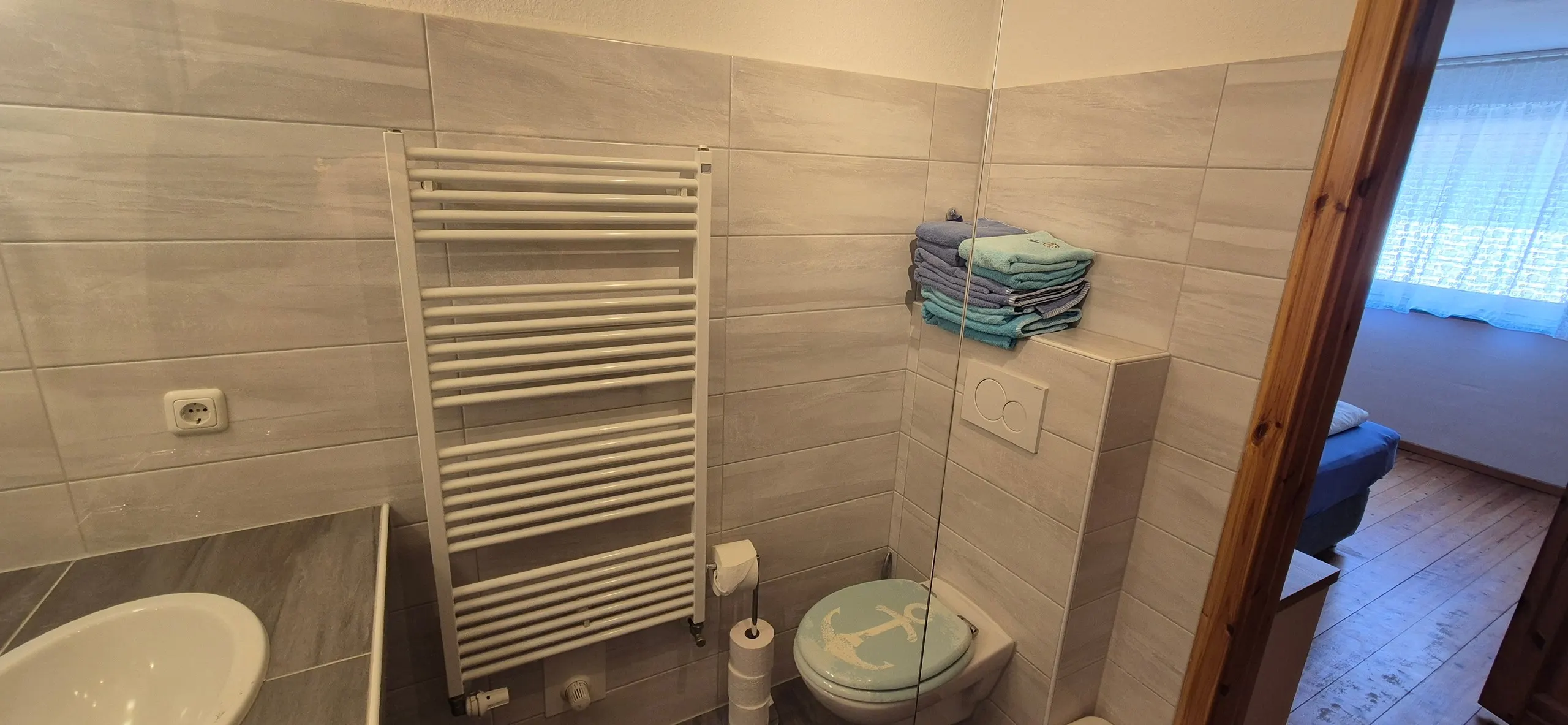 Enzian – Badezimmer mit hellgrauen Fliesen, wandhängender Toilette mit maritimem Anker-Deckel und weißem Handtuch-Heizkörper mit …