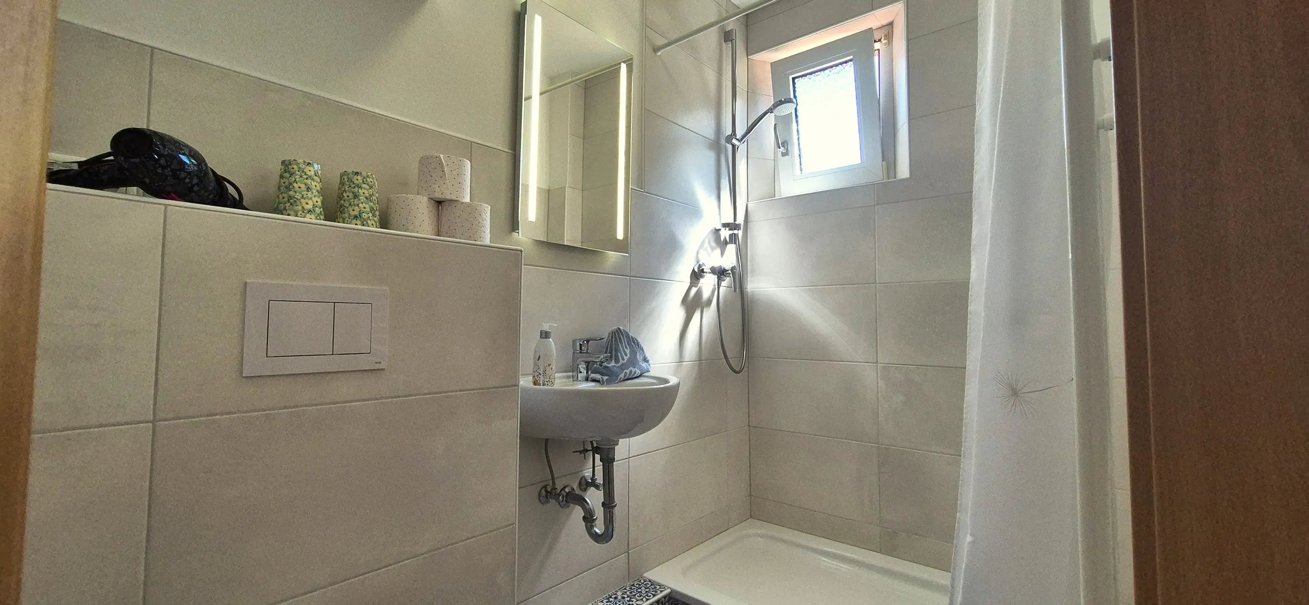 Tanzmaus – Schmales Badezimmer mit gefliester Dusche mit Duschvorhang, wandhängendem Waschbecken und beleuchtetem Spiegelschrank …