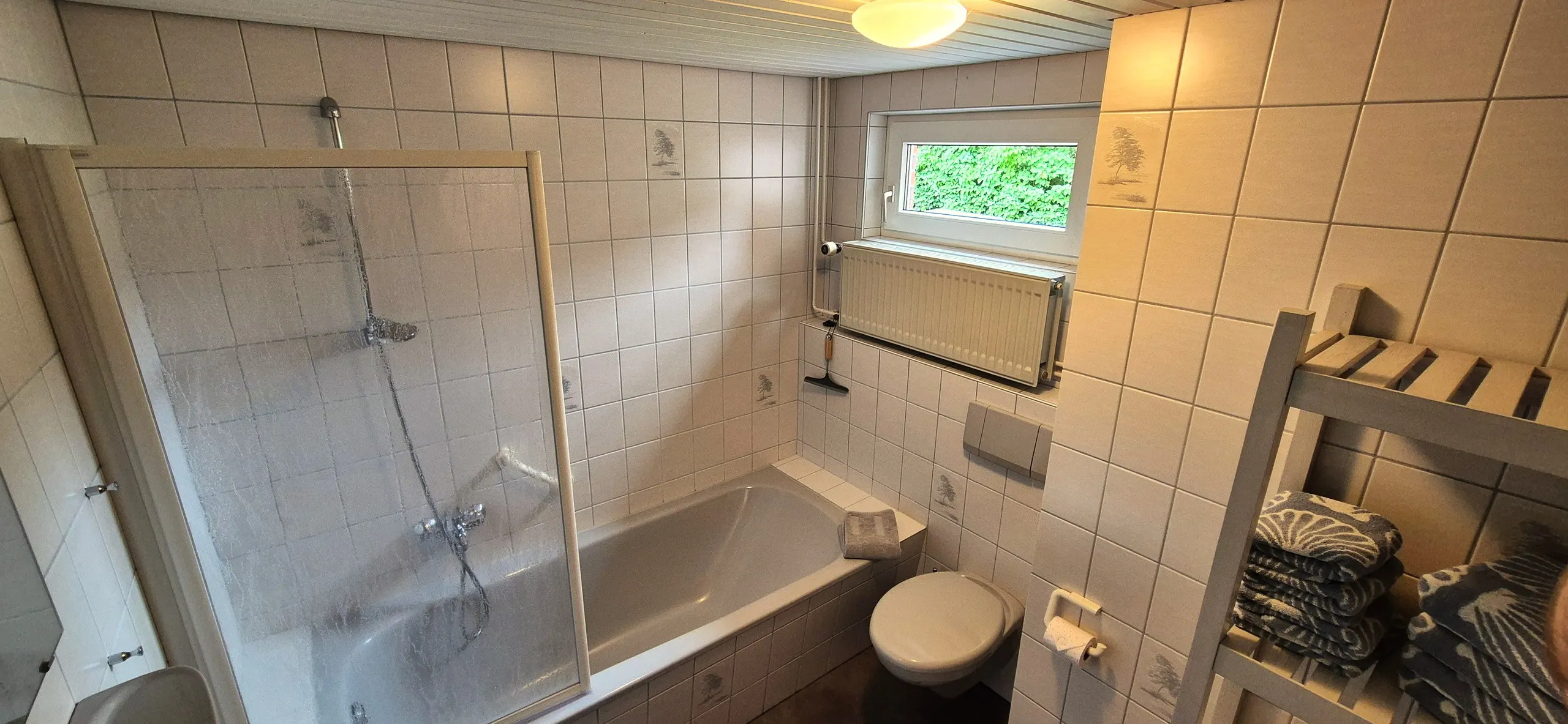 Spitzmaus – Gefliestes Badezimmer mit Duschkabine über der Badewanne, WC und Fenster mit Blick ins Grüne.