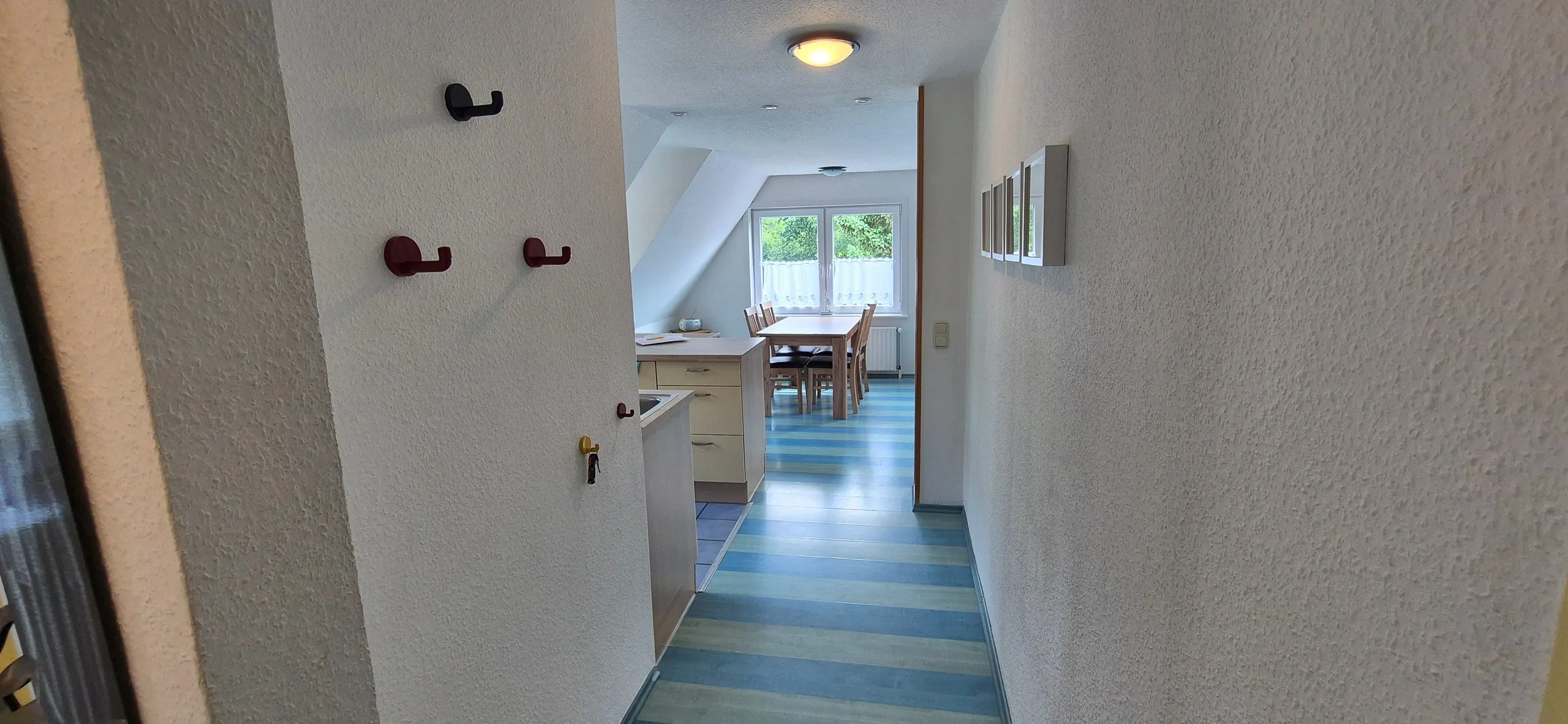 Dünenrose – Schmaler Flur mit weißen und dunkelroten Garderobenhaken an der linken Wand sowie einer Bilderleiste mit vier kleinen …