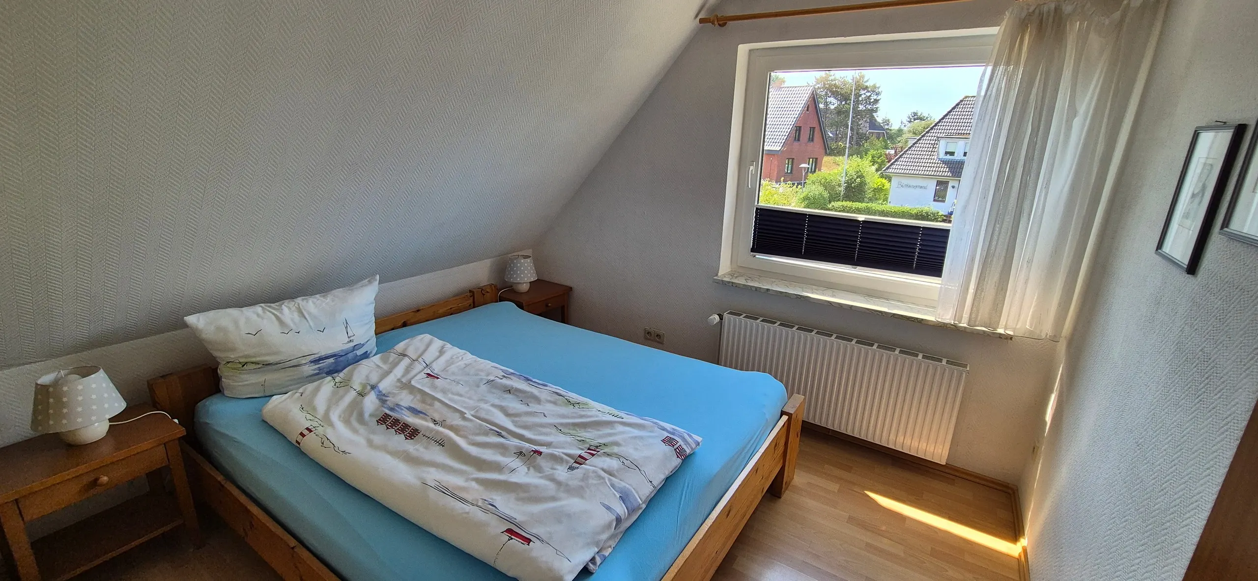 Wollgras – Schlafzimmer mit Doppelbett aus hellem Holz, hellblauer Bettwäsche mit maritimem Leuchtturm-Motiv, links und rechts vom …