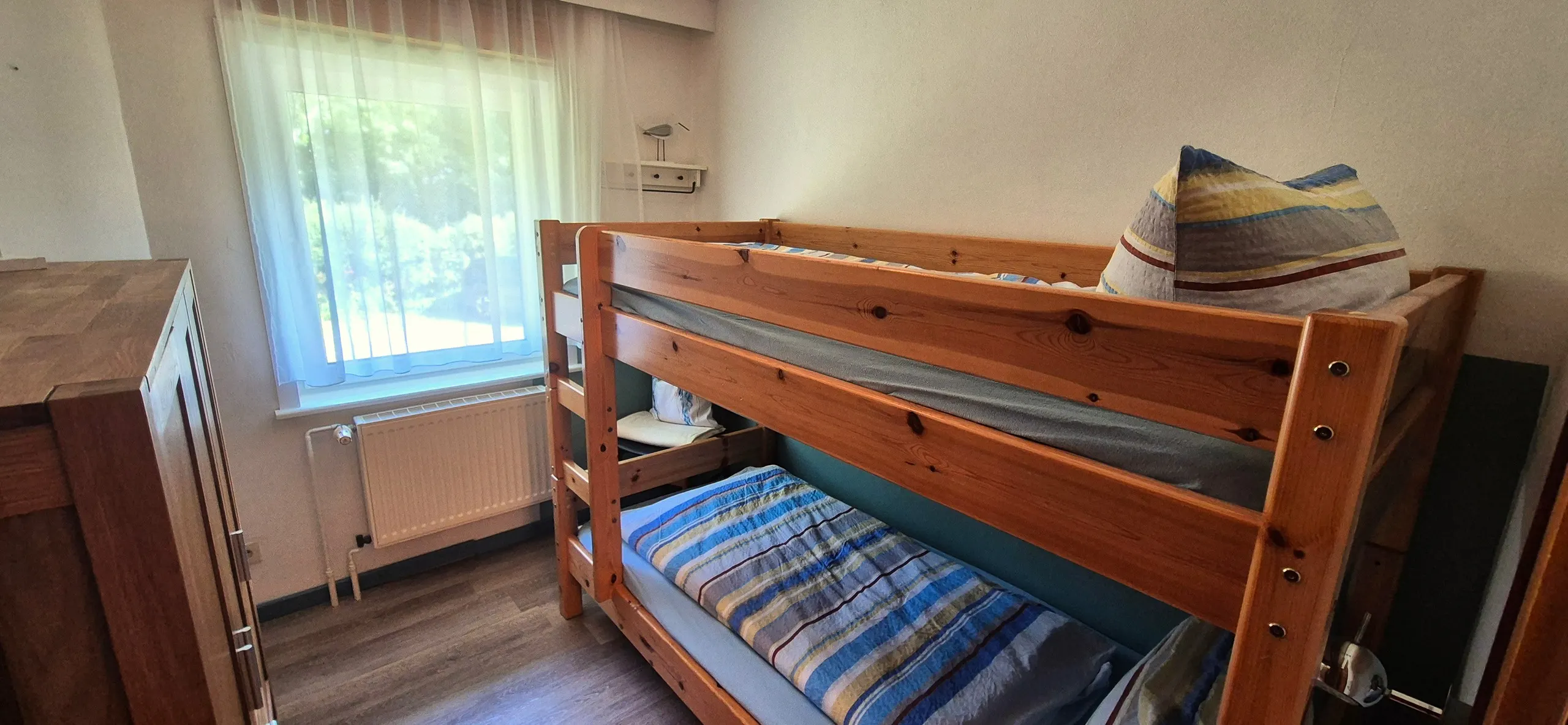 Butt – Zweites Schlafzimmer mit Etagenbett aus Holz, gestreifter Bettwäsche in Blautönen und Blick durch das Fenster ins Grüne.