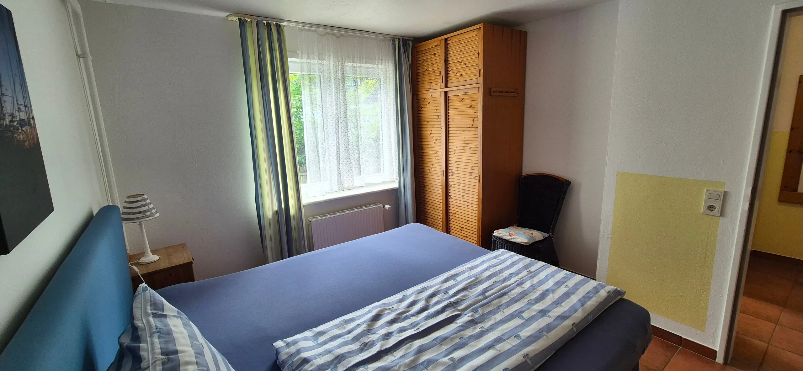 Strandflieder – Schlafzimmer mit dunkelblauem Polsterbett und gestreifter Bettwäsche, davor ein Kiefernholz-Kleiderschrank mit …