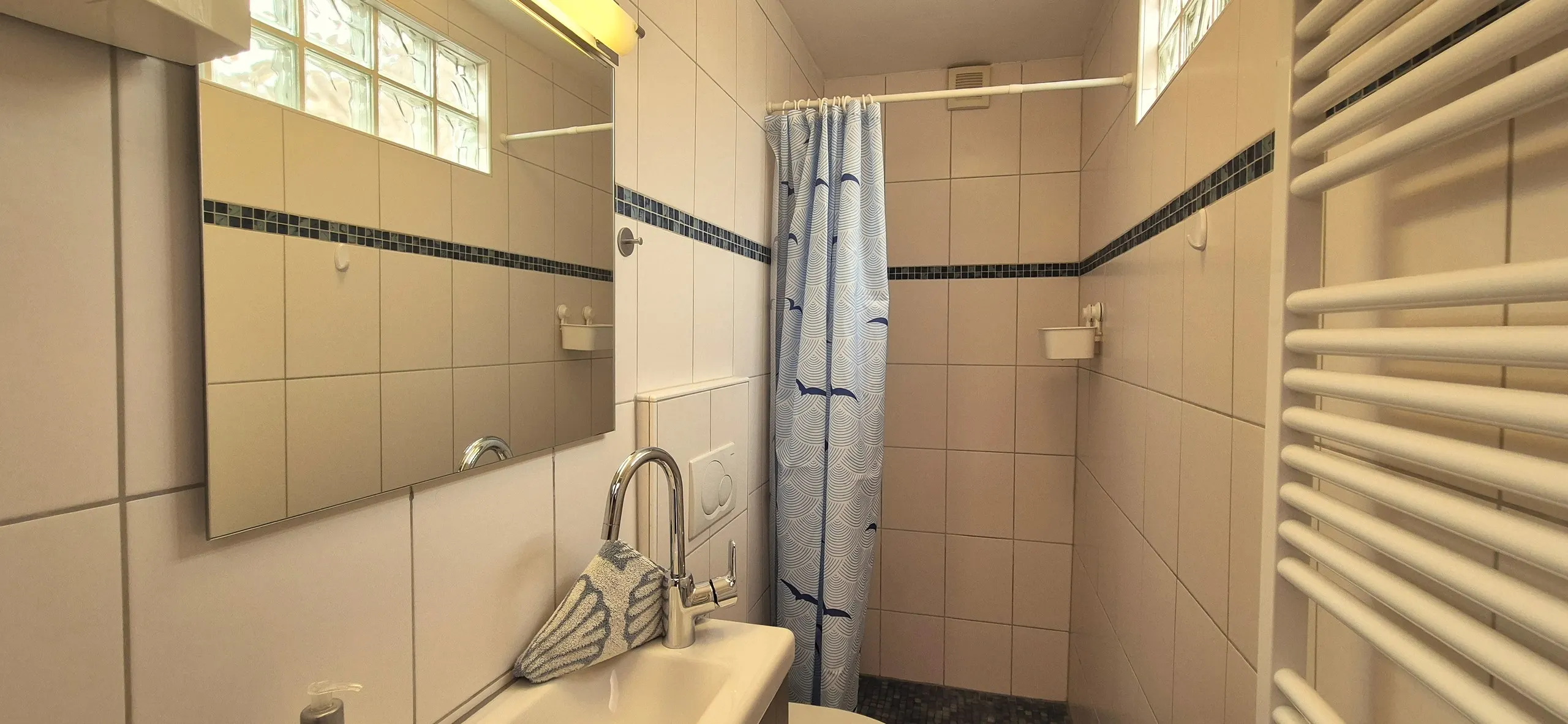 Feldmaus – Kleines Badezimmer mit hellen Wandfliesen und dunklem Mosaik-Fliesenband.