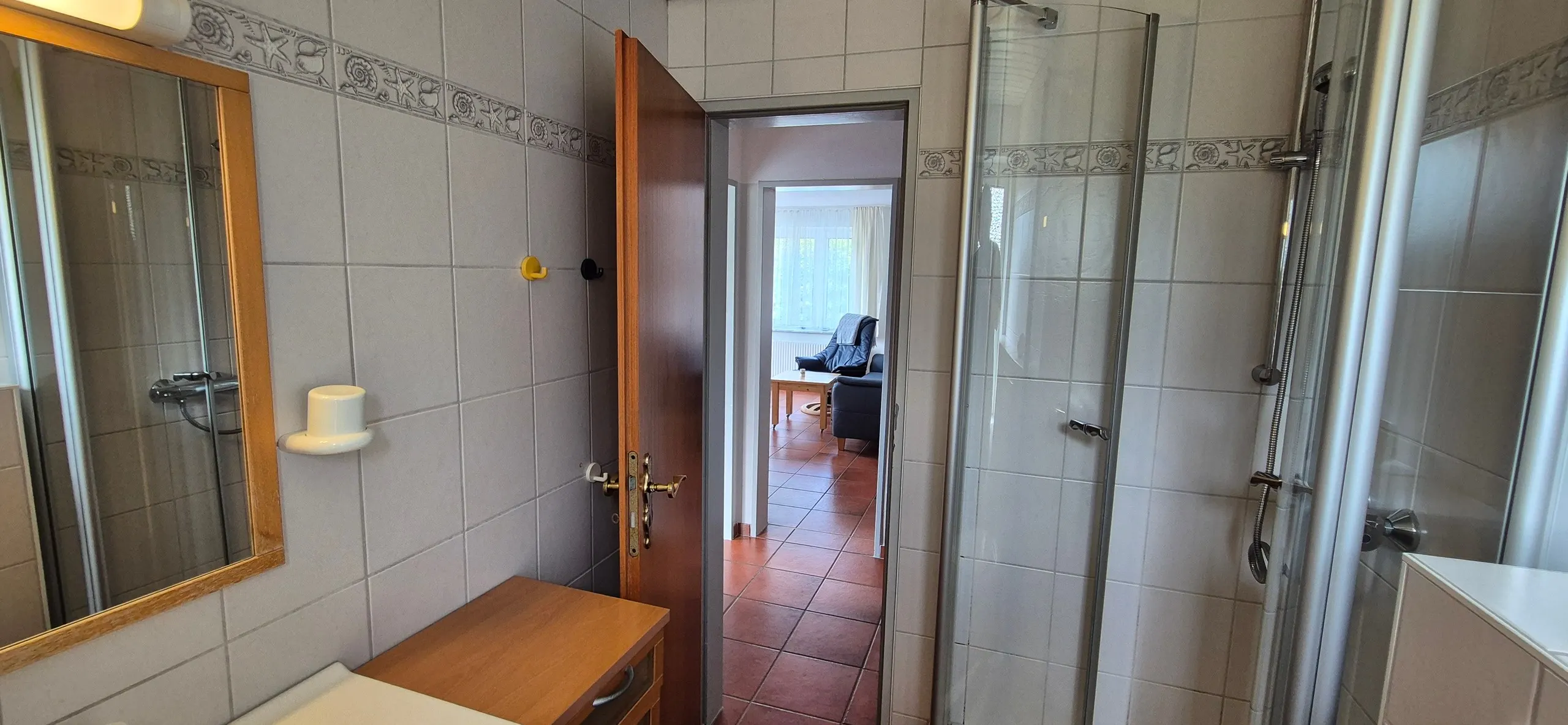 Sonnentau – Gefliestes Badezimmer mit gläserner Duschkabine rechts und einem Wandspiegel mit Holzrahmen über einer Holzkommode …