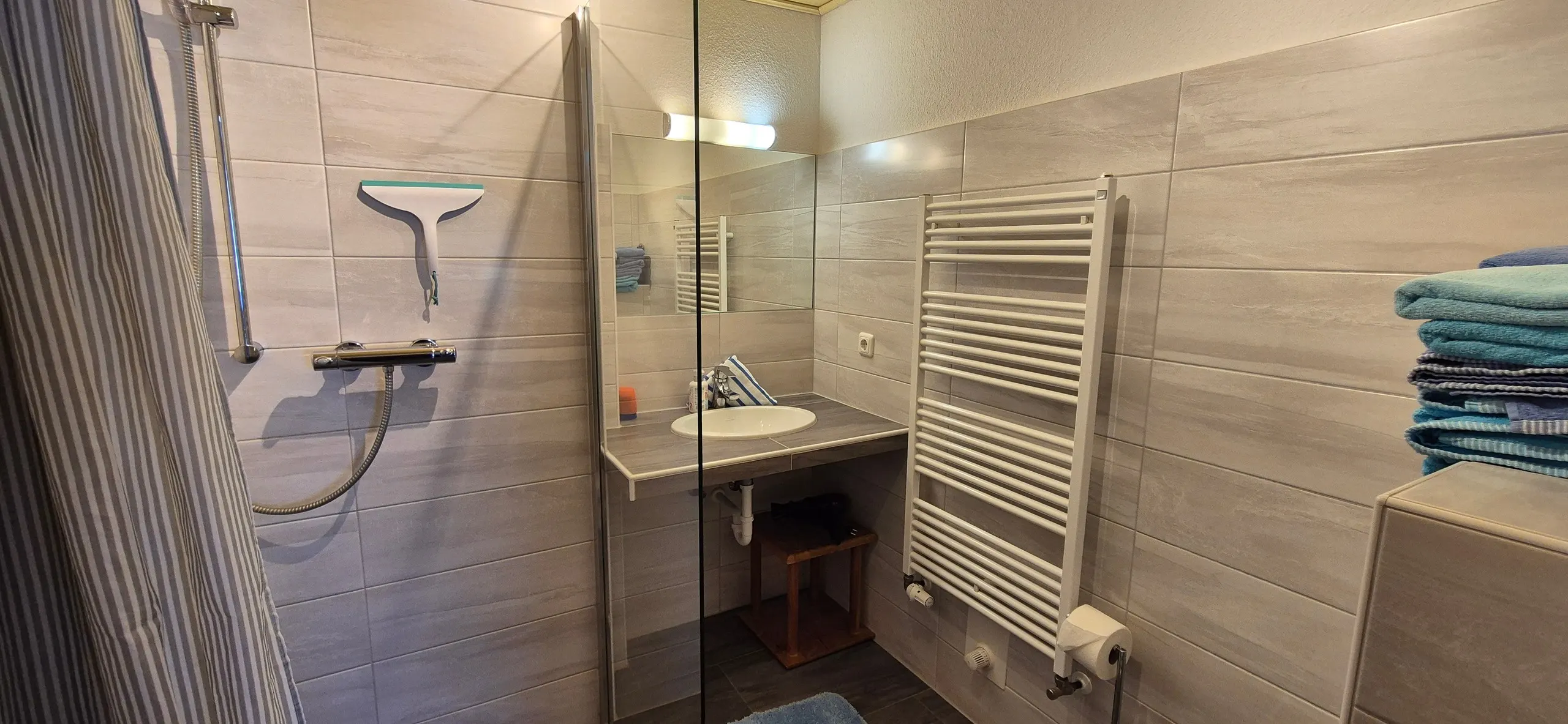 Enzian – Badezimmer mit begehbarer Glas-Duschkabine, Thermostat-Armatur und Abzieher an der Fliesenwand links.