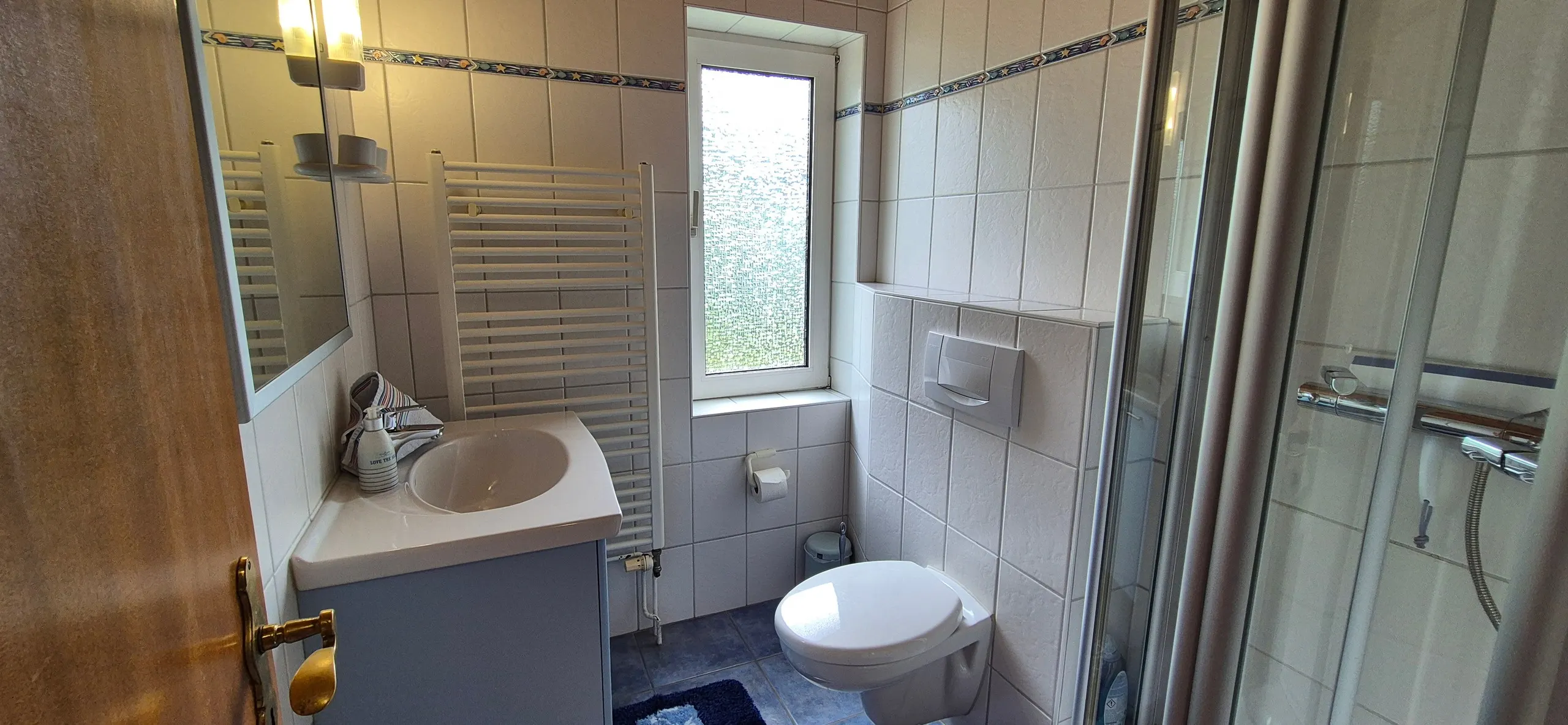 Strandflieder – Badezimmer mit weißen Wandfliesen, blauer Sternchen-Bordüre und einem weißen Handtuchheizkörper unter dem Fenster mit …