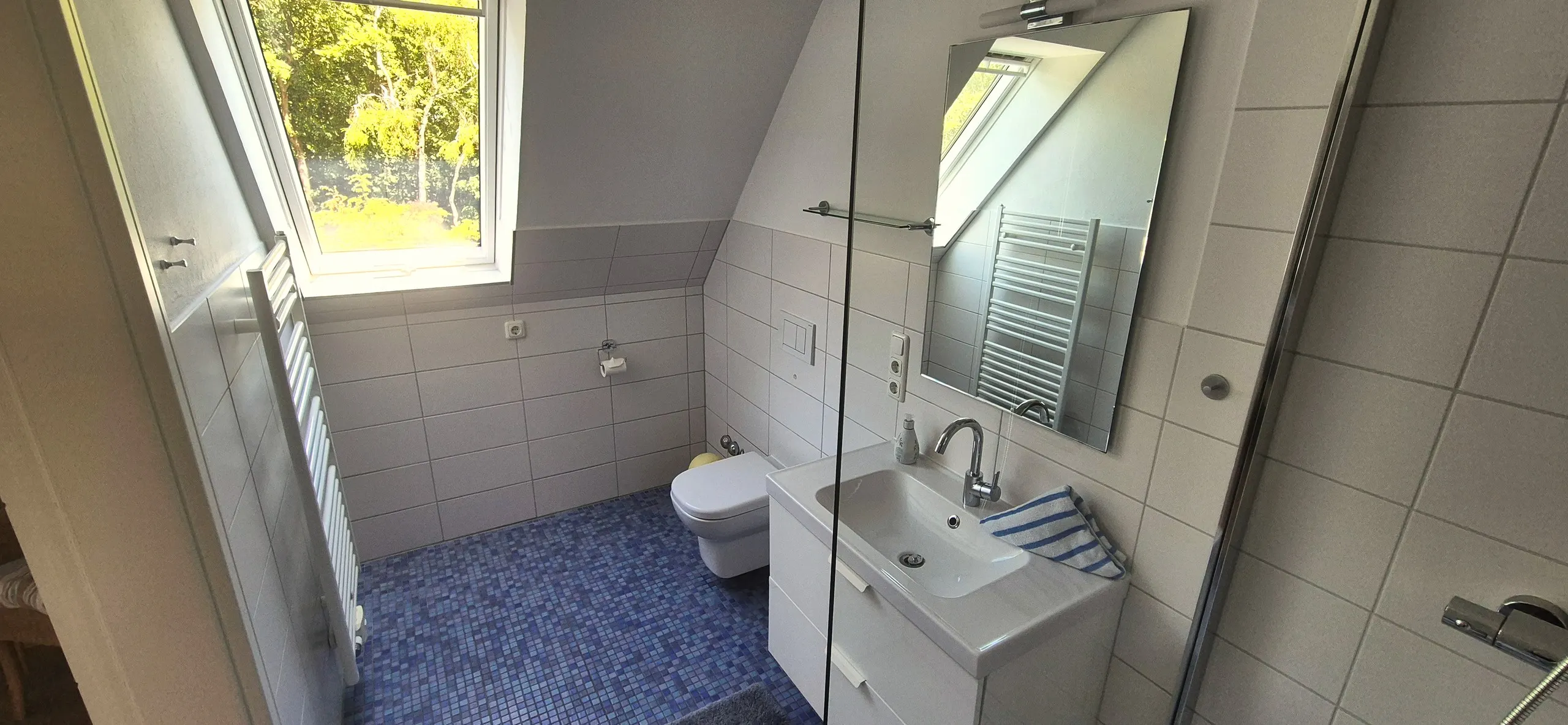 Seestern – Badezimmer unter der Dachschräge mit Dachflächenfenster und Blick ins Grüne, blauem Mosaikfliesenboden und hellgrauen …