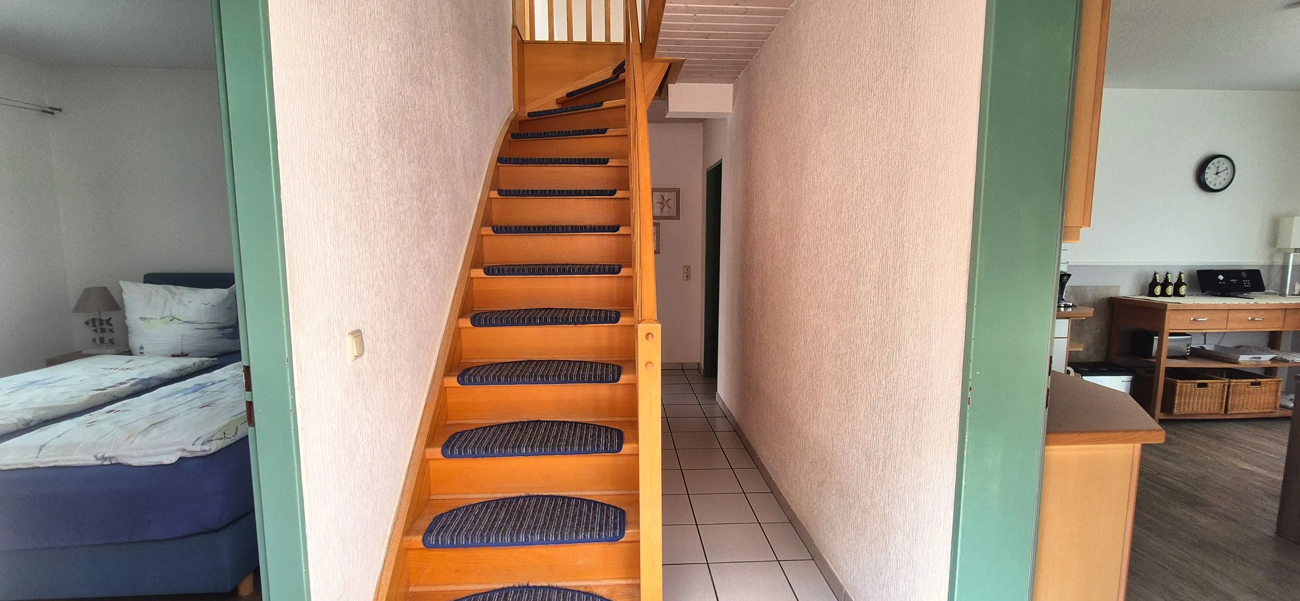 Küstenhaus – Zentraler Flurbereich mit hölzerner Treppe hoch ins Obergeschoss, die Stufen sind mit blauen Stufenmatten belegt.