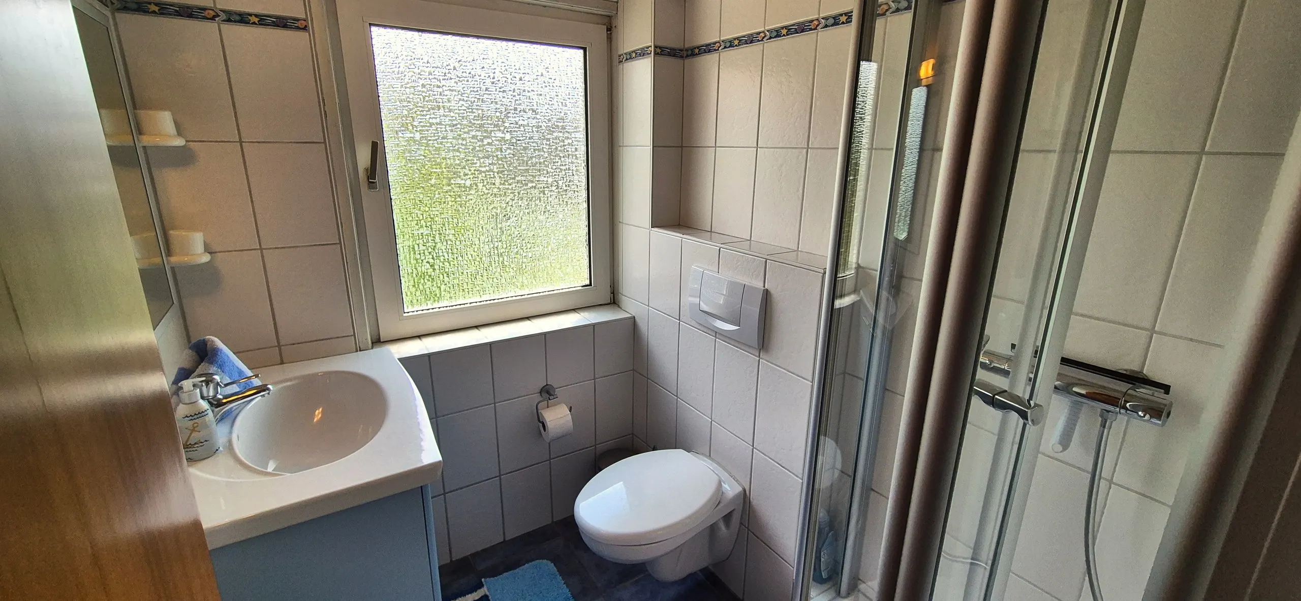 Glockenblume – Badezimmer mit weißen Wandfliesen und blauem Bordürenband, bodengleicher Duschkabine mit Glastür und Chrom-Duscharmatur …