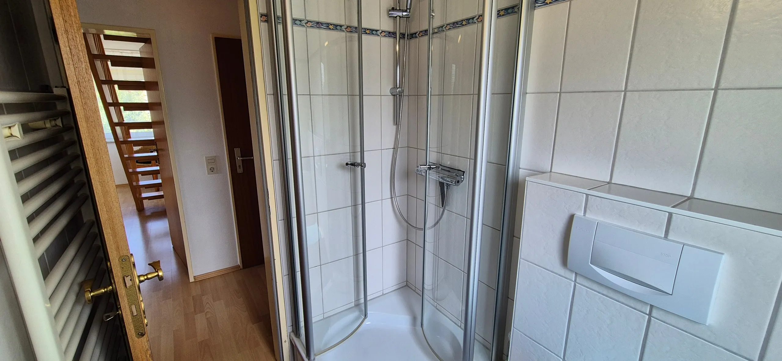 Wollgras – Blick in ein gefliestes Badezimmer mit halbrunder Glas-Eckdusche und Regendusch-Armatur, weiße Fliesen mit blauem …
