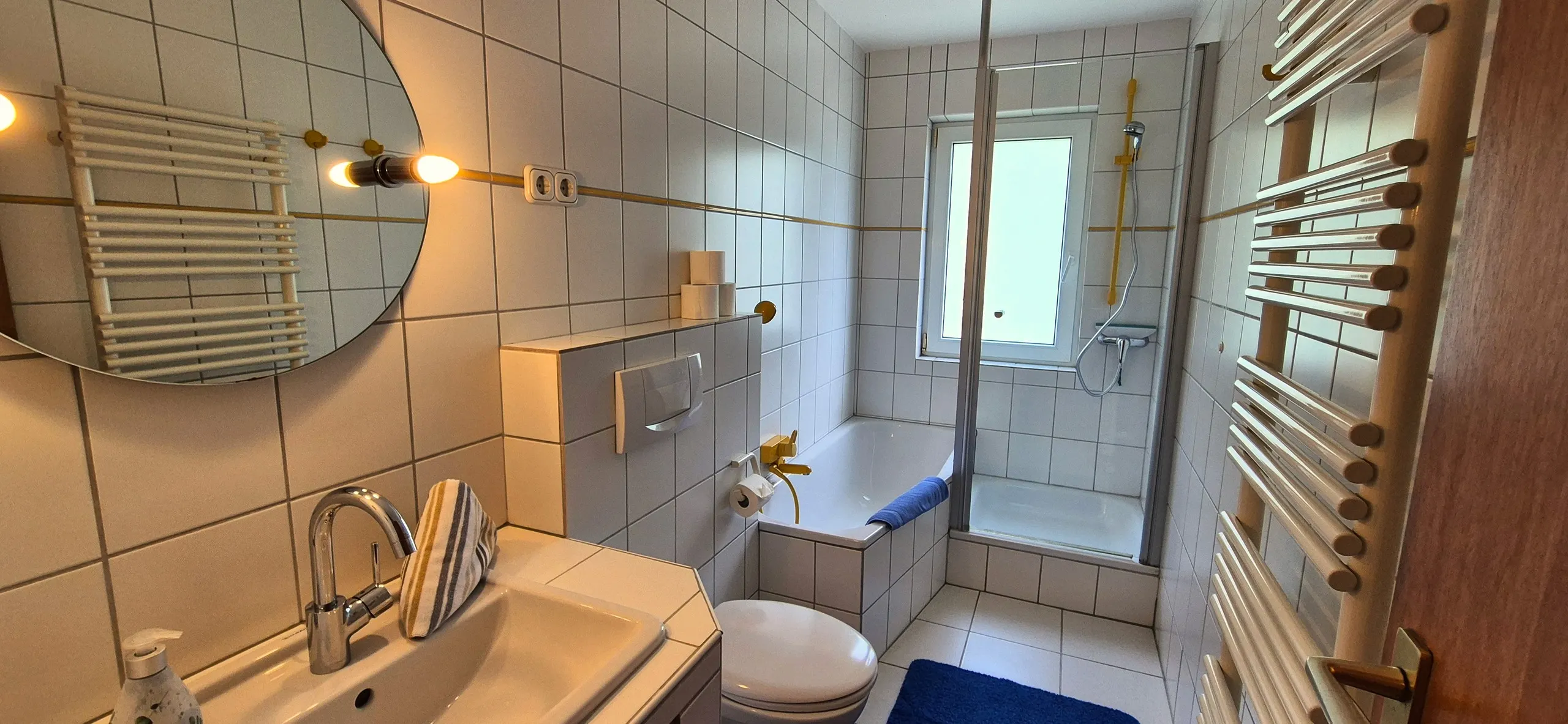 Austernfischer – Gekacheltes Badezimmer mit Badewanne und separater Duschkabine, rundem Spiegel über dem Waschbecken und einem …