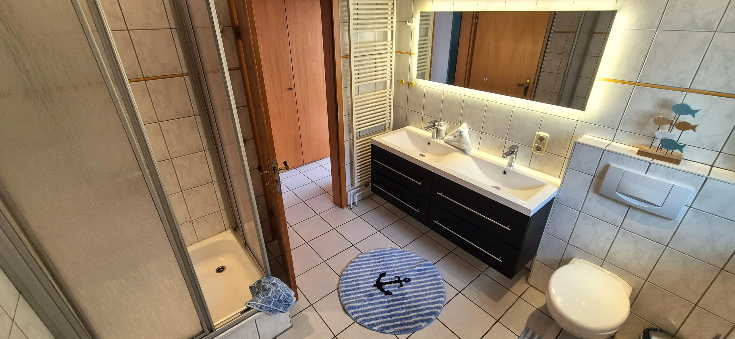 Küstenhaus – Geradliniges, modernes Badezimmer mit heller Fliesenoptik in Marmor-Look und goldenen Zierstreifen.