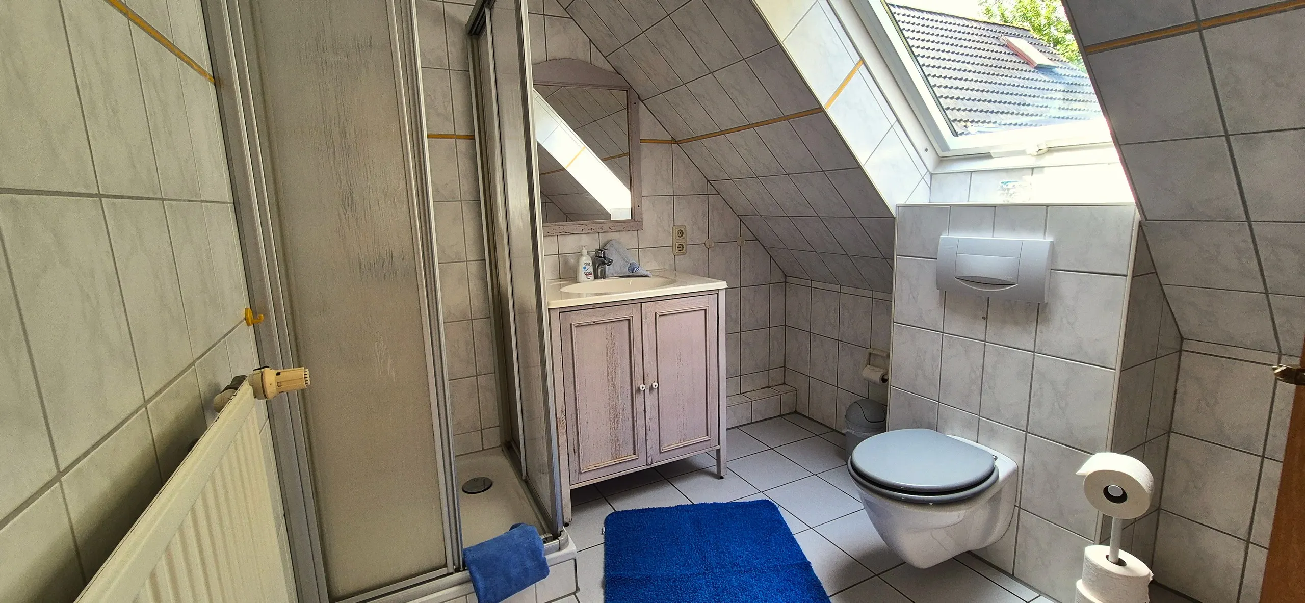 Küstenhaus – Badezimmer im Dachgeschoss mit heller Fliesung und gelben Zierstreifen, geprägt durch die schräge Wand mit großem …