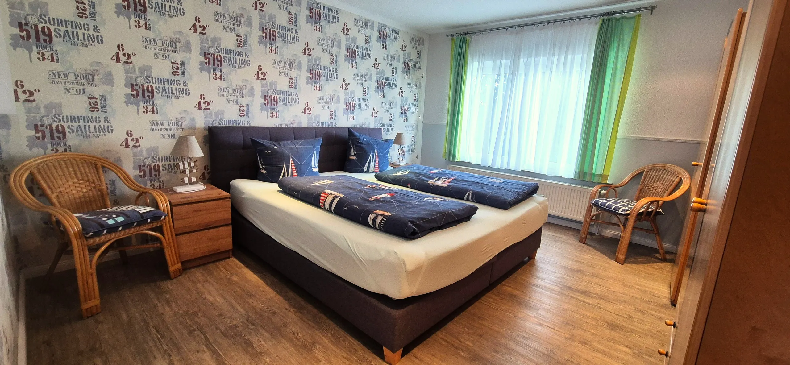 Küstenhaus – Doppelzimmer mit auffälliger Tapete im Segel-/Vintage-Schriftzugdesign in Blau-Rot-Tönen an der Bettwand.