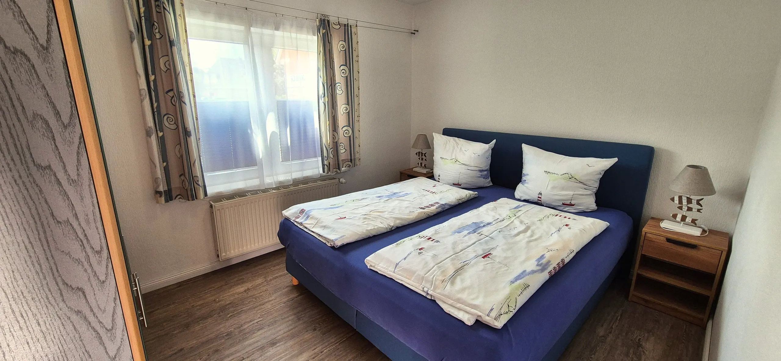 Küstenhaus – Schlichtes, helles Schlafzimmer mit Doppelbett mit dunkelblauem Polsterkopfteil und passender Bettwäsche mit …